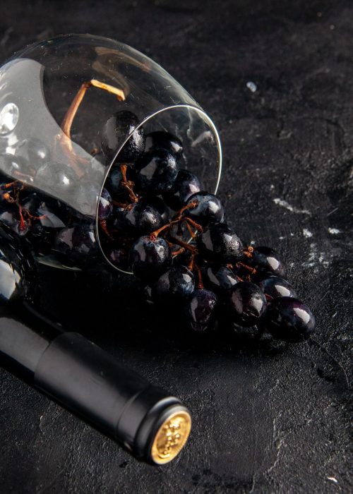 bottom-view-black-grapes-overturned-wine-glass-wine-bottle-dark-table-min